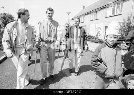 Stadtrat Van Veelen betrachtet die Wanderroute für Kinder in Velserbroek Velserbroek Aldermen Velserbroek, Whizgle News, Dutch Desk, Niederlande, 1950 - 2000 am 12-05-1989. Dies sind die Elemente im Bild. Die Szene zeigt eine lebhafte Straßenlage an einem sonnigen Tag mit einer Mischung aus fröhlichen und informellen Interaktionen unter einer kleinen Gruppe von Menschen. Im Vordergrund laufen drei Erwachsene nebeneinander, jeder trägt legere Kleidung. Zwei von ihnen tragen leichte Jacken, eine in einer hellen Farbe und die andere in einem dunkleren Ton, während der dritte Erwachsene in einem leichten B ist Stockfoto