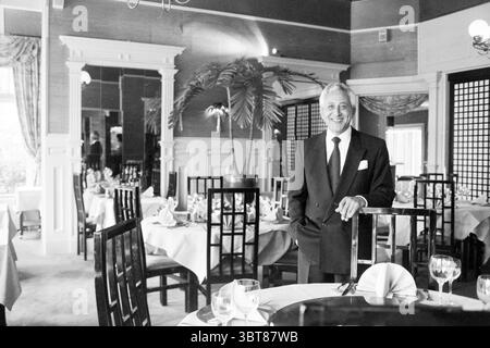 Mr. Shi Amazing Asia Overveen Overveen, Whizgle News, Dutch Desk, Niederlande, 1950 - 2000 am 14-10-1996. Dies sind die Elemente im Bild. Die Szene fängt ein elegantes Interieur des Restaurants ein, das sich durch ein elegantes Ambiente auszeichnet. Der Raum ist mit feinen Details geschmückt, darunter weiche Stühle und Tische, die elegant mit weißen Tischdecken besetzt sind. Jeder Tisch ist mit polierten Glaswaren ausgestattet und einsatzbereit. Im Vordergrund steht ein gut gekleideter Mann selbstbewusst neben einem Stuhl, der einen klassischen schwarzen Anzug mit einem knusprigen weißen Hemd und einer gemusterten Krawatte trägt. Sein Ausdruck ist warm Stockfoto