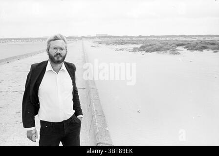 Alderman Kleywegt auf dem IJmuiden Pier Piers and Pier Construction Aldermen IJmuiden the Netherlands, Whizgle News, Dutch Desk, Niederlande, 1950 - 2000 am 30-08-1984. Diese Themen sind in der Abbildung dargestellt. Ein Mann steht zuversichtlich am Rand eines Küstenweges, eingerahmt vor dem Hintergrund eines ruhigen Meeres. Er trägt einen leicht zerzausten Bart und trägt einen dunklen Blazer über einem knusprigen, weißen Hemd, der einen auffälligen Kontrast zu den gedämpften Tönen um ihn herum bildet. Die Einstellung erscheint bedeckt, wobei ein weiches, diffuses Licht die Szene umhüllt und das monochromatische Gefühl verbessert. Der Pat Stockfoto