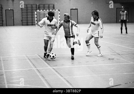 Willems Floet - Rinko Indoor Football, Whizgle News, Dutch Desk, Niederlande, 1950 - 2000 am 15-10-1980. Dies sind die Elemente im Bild. Die Szene fängt einen intensiven Moment in einer kleinen Sporthalle ein, die von einem Gefühl der Dringlichkeit und des Wettkampfes erfüllt ist. Die Spieler tragen kontrastierende Uniformen: Zwei in überwiegend weißer Kleidung mit auffälligen schwarzen Schriftzügen und einer in dunklerem Outfit, das sich durch die helleren Farben auszeichnet. Im Vordergrund dribbelt ein Spieler gekonnt einen Fußball mit konzentrierter Stirn. Er wird auf beiden Seiten von Gegnern flankiert, beide Stockfoto