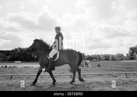 Final NK Iceland Ponies, Whizgle News, Dutch Desk, Niederlande, 1950 - 2000 am 24-07-1993. Diese Themen werden in der Abbildung angezeigt. Die Szene zeigt eine ruhige Reitanlage im Freien, gekennzeichnet durch sanfte, diffuse Beleuchtung, die einen sanften, bewölkten Tag suggeriert. Im Vordergrund steht ein Reiter in formeller Reiterkleidung, bestehend aus einer taillierten Jacke und hohen Reitstiefeln, anmutig auf einem eleganten, dunkelbraunen Pferd. Das Pferd hat eine gut gepflegte Mähne und gleitet auf einem sandigen Pfad mit einem Hauch von Selbstvertrauen. Die Umgebung von Reiter und Pferd bietet eine üppige, grüne Landschaft Stockfoto