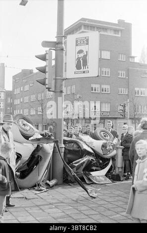 Eine Gruppe von Menschen betrachtet die Wrackteile eines Autos nach einem Verkehrsunfall Amsterdam Vrijheidslaan Niederlande, Whizgle News, Dutch Desk, Niederlande, 1950 - 2000 am 04-1963. Die Abbildung zeigt diese Themen. Die Szene fängt einen Moment des Chaos nach einem Fahrzeugunfall ein. Im Vordergrund liegt ein beschädigtes Auto, dessen Dach gegen den Bürgersteig gepresst ist, wobei verschiedene freiliegende Metallteile und zersplittertes Glas verstreut sind. Der Lack des Fahrzeugs erscheint stumpf, möglicherweise aufgrund von Alter oder Verschleiß, und Schmutz deutet auf die Kraft des Aufpralls hin. Um das Auto herum hat eine Gruppe von Zuschauern Gangart Stockfoto