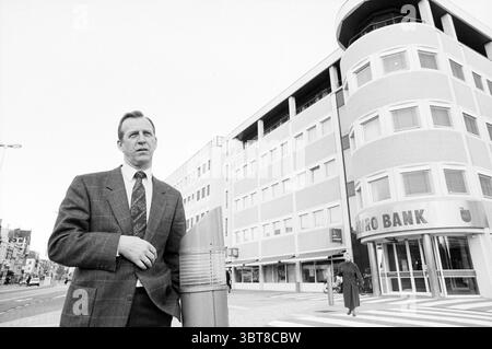 Mann vor dem Amrobank Büro Houtplein Haarlem Houtplein Niederlande, Whizgle News, Dutch Desk, Niederlande, 1950 - 2000 am 16. 02.1990. Diese Themen sind in der Abbildung dargestellt. Die Szene entfaltet sich in einer urbanen Umgebung, in der ein gut gekleideter Mann im Vordergrund steht. Er trägt einen klassischen, maßgeschneiderten Anzug mit einem markanten Karomuster, das eine subtile Textur verleiht. Seine Haltung ist entspannt und dennoch selbstbewusst, wobei eine Hand beiläufig an einem nahe gelegenen Stab ruht. Im Hintergrund hebt sich ein modernes, mehrstöckiges Gebäude hervor, das sich durch sein glattes, rechteckiges Gebäude auszeichnet Stockfoto