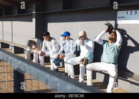 Verschiedene männliche Baseballkameraden sitzen auf der Bank in einer Dugout und halten Fledermäuse und Handschuhe Stockfoto