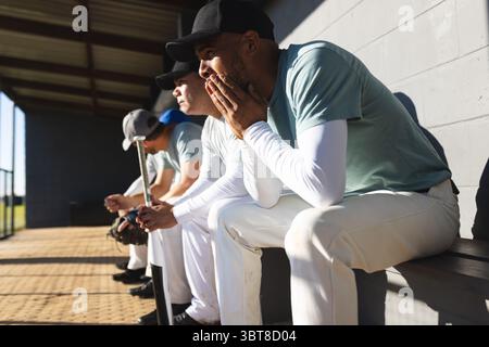 Verschiedene männliche Baseballkameraden in Uniformen sitzen auf der Bank im Dugout und halten Schläger und Handschuhe Stockfoto