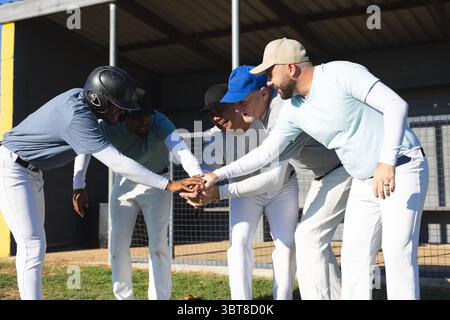Verschiedene männliche Baseballkameraden, die sich von den Dugout-Spielern in die enge lehnen und Hände mit Helm und Kappen stapeln Stockfoto