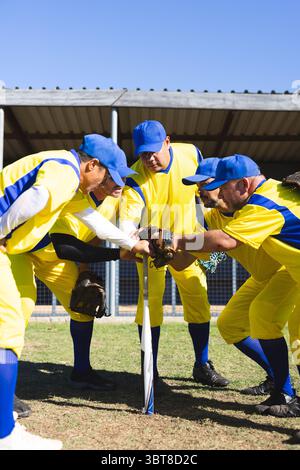 Verschiedene männliche Mannschaftskameraden, die sich in gelben Trikots und Handschuhen um gepflanzte Fledermaus auf dem Grasfeld herumtummeln Stockfoto
