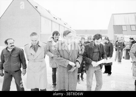 Min. Van Ardenne eröffnet Energiegarten Hoofddorp Hoofddorp the Netherlands, Whizgle News, Dutch Desk, Niederlande, 1950 - 2000 am 14. 01.1985. Dies sind die Themen im Bild. Die Szene fängt eine Gruppe von Individuen ein, die sich durch eine krasse, winterliche Landschaft bewegen, die von einer überwältigenden weißen und grauen Palette gekennzeichnet ist. Weiches, diffuses Licht bedeckt den Bereich und deutet auf bewölkten Himmel hin, der zu einer gedämpften Atmosphäre beiträgt. Im Vordergrund gehen mehrere Männer eng zusammen, ihre Ausdrucksweisen variieren – einige erscheinen ernst oder kontemplativ, während andere Anzeichen von Unwohlsein oder Müdigkeit zeigen. Sie sind b Stockfoto