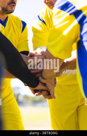 Verschiedene männliche Teamkollegen tragen gelbe Trikots, die sich bei Sonnenlicht auf dem Sportplatz die Hände stapeln Stockfoto