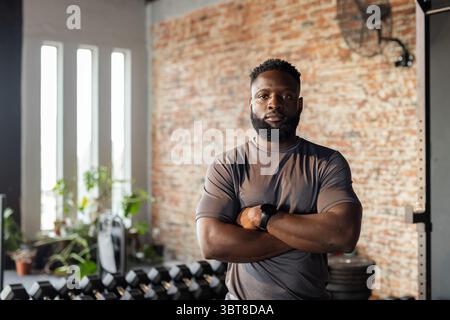 Afroamerikanischer Fitnesstrainer mit Sportbekleidung, der Arme im Fitnessstudio am Hantelständer kreuzt Stockfoto