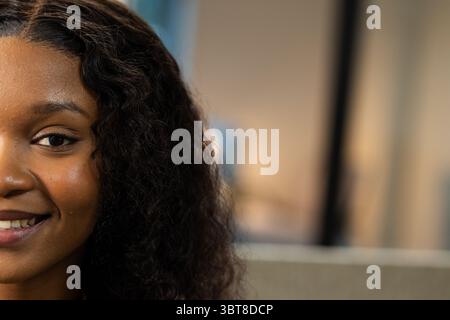 Afroamerikanerin lächelt leise im Innenraum in der Nähe von Fensterrahmen in warmem Licht, Kopierraum Stockfoto