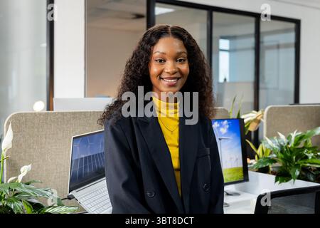 Afroamerikanerin steht und lächelt im Großraumbüro am Schreibtisch mit Laptop-Monitoranlagen Stockfoto