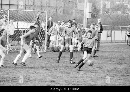 Schulfußball Fulmars, Whizgle News, Dutch Desk, Niederlande, 1950 - 2000 am 12-04-1993. Diese Themen sind in der Abbildung dargestellt. Die Szene zeigt ein lebhaftes Jugendfußballspiel auf einem grasbewachsenen Feld, das von einer energiegeladenen Atmosphäre geprägt ist. Im Vordergrund spielen zwei Spieler: Der eine dribbelt einen Fußball, der andere verfolgt den Wettkampf. Sie sind beide in sportlicher Freizeitausrüstung gekleidet, überwiegend in Grau- und Schwarztönen, die mit den monochromatischen Tönen des Gesamtbildes harmonieren. Hinter ihnen befindet sich eine Gruppe von Zuschauern, bestehend aus mehreren Stockfoto