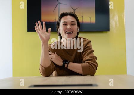 Asiatischer Mann, der mit der Hand winkt und lächelt, während er am Tisch im Büro mit Windturbinen-Display sitzt Stockfoto