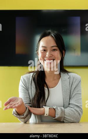 Asiatische Frau, die die linke Hand ausstreckt und lächelt, mit Smartwatch am Schreibtisch vor dem Bildschirm Stockfoto