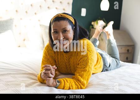 Afroamerikanerin, die sich auf dem Bett liegend mit einem Kopfband aus Senfpullover gegen ein getuftetes Kopfteil trägt Stockfoto