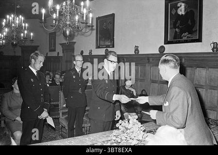 Zandvoort Fire Department Award Fire Department, Whizgle News, Dutch Desk, Niederlande, 1950 - 2000 am 03-06-1969. Das Bild enthält diese Themen. Die Szene spielt in einem schwach beleuchteten Raum mit üppiger Holztäfelung, die ein Gefühl von Geschichte und Formalität verleiht. Der Raum wird durch warmes Licht von zahlreichen Kronleuchtern beleuchtet, die von der Decke hängen. Jeder von ihnen ist mit mehreren Kerzen geschmückt, die sanft flackern und eine weiche, einladende Atmosphäre schaffen. Im Vordergrund steht ein Mann in einer dunklen Uniform mit einem Abzeichen mit einem feierlichen Ausdruck, der ein Dokument eines anderen Mannes annimmt Stockfoto