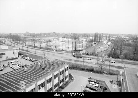Übersicht der GEB-Website, Whizgle News, Dutch Desk, Niederlande, 1950 - 2000 am 06-02-1992. Dies sind die Elemente im Bild. Die Szene zeigt eine weitläufige urbane Landschaft, die von einem erhöhten Aussichtspunkt aus gesehen wird. Im Vordergrund dominiert ein großes Gebäude mit flachem Dach den unteren Teil des Anblicks mit einem Muster aus dunklen Fliesen, die subtile Variationen zeigen. Das Äußere des Gebäudes ist meist hell, vielleicht ein blassgraues oder weißes Licht, das sich von dem dunkleren Dach abhebt. Direkt hinter dem Gebäude verläuft eine Straße am unteren Rand des Bildes, gesäumt von geparkten Fahrzeugen in geordneten Reihen. T Stockfoto