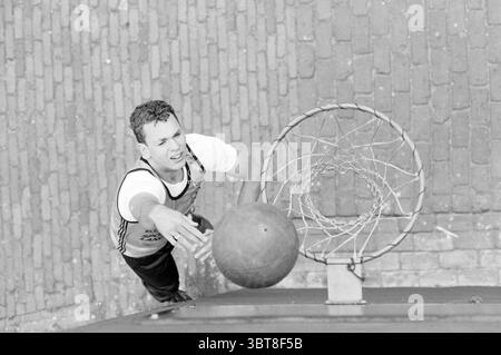 Basketballspieler, Whizgle News, Dutch Desk, Niederlande, 1950 - 2000 am 06-1988. Dies sind die Elemente im Bild. In einer dynamischen Umgebung im Freien wird ein junger Mann während der Action gefangen, während er versucht, einen Basketballschuss zu machen. Er befindet sich direkt unter einem Basketballkorb, der von Rand und Netz markant eingerahmt wird. Der Athlet trägt ein helles ärmelloses Oberteil und dunkle Shorts, was seinen sportlichen Körper verbessert und Bewegung suggeriert. Sein Ausdruck ist fokussiert und zeigt Konzentration, während er mit einem ausgestreckten Arm in Richtung des Basketballs reicht Stockfoto