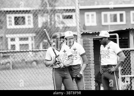 Softball Kennemerland - Antillian Stars 28-06-1991, Whizgle News, Dutch Desk, Niederlande, 1950 - 2000 am 28-06-1991. Diese Themen werden in der Abbildung angezeigt. Eine Gruppe von drei Spielern steht in der Nähe eines Maschendrahtzaugs, der ein Baseballfeld umschließt. Im Vordergrund liegt ein junger Mann mit einem Fledermaus auf der Schulter, der einen Helm und ein Trikot trägt, das mit einem Sternendesign und dem Namen „Caribbean Stars“ verziert ist. Sein Ausdruck ist fokussiert und zeigt tiefe Konzentration oder Vorfreude an. Neben ihm lehnt sich ein etwas älterer Mann in ähnlicher Kleidung lässig mit überkreuzten Armen an den Zaun. Er hat ein c Stockfoto
