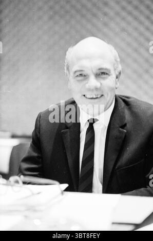 Portraits of council Members., Whizgle News, Dutch Desk, Niederlande, 1950 - 2000. Das Bild enthält diese Themen. Die Szene zeigt einen Mann, der an einem Tisch sitzt und ein warmes und nahes Auftreten ausstrahlt. Er hat einen glatten Glatzkopf und ein freundliches Lächeln, das seinen Charakter zum Ausdruck bringt. Er trägt einen klassischen schwarzen Anzug mit einem knusprigen weißen Hemd und trägt eine dunkle Krawatte mit dezenten Streifen, die seinem Aussehen einen Hauch von Eleganz verleiht. Der Hintergrund zeigt eine strukturierte Wand, die wahrscheinlich ein sich wiederholendes Muster aufweist, das der Einstellung Tiefe verleiht. Sanfte, diffuse Beleuchtung hebt die Eigenschaften des Mannes hervor, Crea Stockfoto