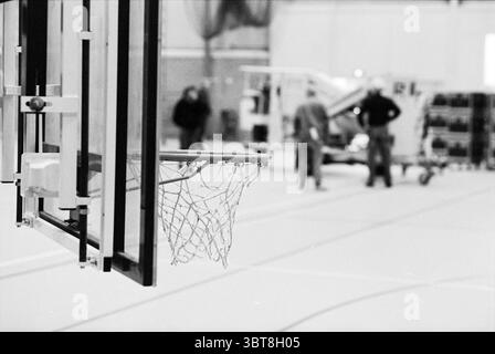 Bau der CIOS-Halle, Whizgle News, Dutch Desk, Niederlande, 1950 - 2000 am 21-12-1993. Dies sind die Themen im Bild. In einem geräumigen Fitnessraum befindet sich im Vordergrund ein Basketballkorb, dessen Netz leicht wiegt. Der Metallrahmen des Rings leuchtet unter den Deckenleuchten, die einen sanften Glanz über den polierten Holzboden werfen. Der Hintergrund zeigt zwei Figuren, die in Gesprächen involviert sind. Ihre Silhouetten sind leicht verschwommen, was auf ein Gefühl von Tiefe und Distanz hindeutet. Die Farben der Szene sind überwiegend gedämpft, mit Grautönen und subtilen Anklängen von Wärme aus dem Holzboden und dem Th Stockfoto