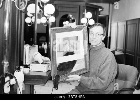 Mr. Carpenter + Painting H'lem Haarlem the Netherlands, Whizgle News, Dutch Desk, Niederlande, 1950 - 2000 am 27-02-1996. Diese Themen sind in der Abbildung dargestellt. Die Szene zeigt einen Mann, der in einem gut beleuchteten Innenraum sitzt und sich durch eine Mischung aus modernen und klassischen Elementen auszeichnet. Er hält ein gerahmtes Kunstwerk, das eine Nahaufnahme des Gesichts einer Frau mit langen, fließenden Haaren zeigt, wahrscheinlich ein auffälliges zentrales Thema des Stücks. Der Rahmen selbst ist verziert und weist auf einen Hauch von Eleganz hin. Der Mann hat einen ziemlich ernsten Ausdruck und seine Kleidung ist lässig, bestehend aus einem Pullover. Seine Brille gibt Stockfoto