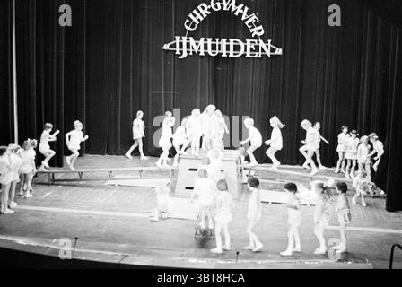 Aufführung des Christian Gymnastics Association 'IJmuiden' auf der Bühne eines Theaters., Whizgle News, Dutch Desk, Niederlande, 1950 - 2000. Dies sind die Themen im Bild. Die Szene zeigt eine lebhafte Bühnenvorstellung, die in einer Theaterumgebung stattfindet. Die Bühne ist voll von zahlreichen Kindern, die alle in schlichten, hellen Outfits gekleidet sind, was eine sorglose, verspielte Atmosphäre suggeriert. Ihre Jugendliche Energie ist spürbar, mit einigen Momenten animierter Bewegung – tanzen, springen und auf der Bühne rennen. Die Kulisse ist mit einem großen, stilisierten Schild geschmückt, das indiziert erscheint Stockfoto