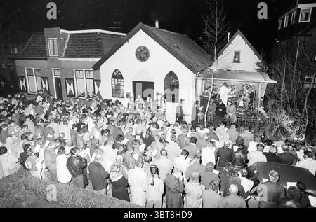 Christmas carol Senf Seed Santpoort Christmas and Christmas Trees Santpoort, Whizgle News, Dutch Desk, Niederlande, 1950 - 2000 am 24-12-1988. Diese Themen sind in der Abbildung dargestellt. In der Szene versammelt sich eine große Menschenmenge vor einem malerischen, bescheidenen Gebäude, das einen besonderen architektonischen Stil aufweist, der durch ein kleines Rosenfenster und ein Giebeldach gekennzeichnet ist. Die Fassade des Gebäudes, wahrscheinlich in einer hellen Farbe, steht im Kontrast zu den tiefen Schatten des Nachthimmels. Die Menge, die sich aus zahlreichen Individuen zusammensetzt, umfasst eine Reihe von Alters- und Geschlechtern, alle in Kleidung der Mitte des 20. Jahrhunderts gekleidet, mit vielen W Stockfoto