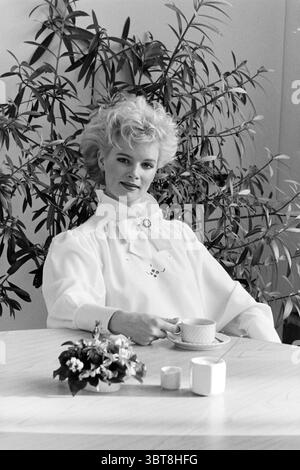 Fashion Report Christmas Fashion, Whizgle News, Dutch Desk, Niederlande, 1950 - 2000 am 06-12-1985. Dies sind die Themen im Bild. In einem sanft beleuchteten Innenbereich sitzt eine Frau anmutig an einem runden Holztisch. Ihre Frisur ist voluminös und elegant gestylt, was ihrem Aussehen einen Hauch von Raffinesse verleiht. Sie trägt eine fließende weiße Bluse mit hohem Kragen, die einen schönen Kontrast zu ihren hellen Details bildet. Um sie herum ist eine üppige grüne Pflanze mit langen, schlanken Blättern, die eine ruhige und einladende Atmosphäre schafft. Die leuchtenden Farben der Pflanze unterstreichen die Einfachheit der Szene. Ein Stockfoto
