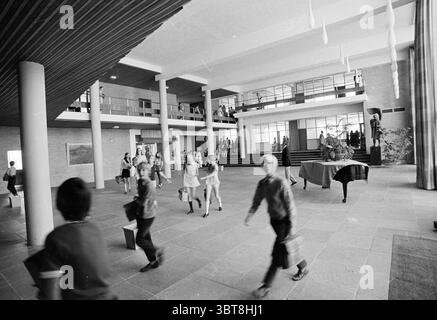 Extra und INT. Sancta Maria Schools Haarlem Van Limburg Stirumstraat Niederlande, Whizgle News, Dutch Desk, Niederlande, 1950 - 2000 am 07-09-1971. Das Bild enthält diese Themen. Die Szene zeigt ein geräumiges, zeitgenössisches Atrium voller Bewegung und Energie. Die Architektur verfügt über hohe Decken, die von großen, weißen zylindrischen Säulen gestützt werden, die dem Raum ein Gefühl von Pracht verleihen. Die Wände sind eine Mischung aus hellen Farbtönen, ergänzt durch Holzakzente, insbesondere die Decke, die eine reiche Textur aufweist, die sich mit den glatten Oberflächen abhebt. Im Vordergrund, mehrere Leute Stockfoto