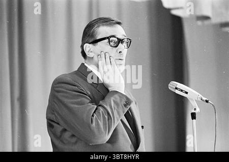 Ria Valk mit Gitarre Andre Meurs Cabaret, Whizgle News, Dutch Desk, Niederlande, 1950 - 2000 am 13.04.1966. Die Abbildung zeigt diese Themen. In einer formellen Umgebung steht ein Mann an einem Mikrofon und strahlt einen Hauch von Nachsicht und Konzentration aus. Er trägt einen maßgeschneiderten Anzug, der dunkel ist und zu einem weißen Hemd und einer Krawatte passt, die einen subtilen Hauch von Raffinesse verleiht. Seine Brille ist dickgerahmt, was zu seinem intellektuellen Verhalten beiträgt. Der Mann scheint mitten in der Rede zu sein, mit einer Hand, die leicht an seiner Wange liegt, was darauf hindeutet, dass er entweder über seine nächsten Worte nachdenkt oder über einen Ser nachdenkt Stockfoto