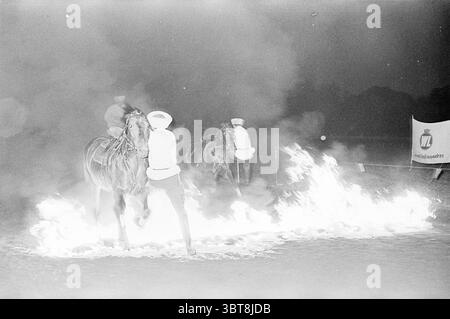 Cossack Riders Groups, Whizgle News, Dutch Desk, Niederlande, 1950 - 2000 am 16-06-1964. Die Abbildung zeigt diese Themen. Die Szene spielt in einer dunklen, nächtlichen Umgebung, mit einer rauchigen Atmosphäre, die einige Details verdeckt und das Drama verstärkt. Zwei Reiter auf dem Pferd stehen im Mittelpunkt der Komposition, ihre Halterungen tauchen aus dem Dunst auf, während sie durch einen Streifen von hellen Flammen navigieren, die auf dem Boden flimmern und tanzen. Die Pferde, stark und muskulös, zeigen Anzeichen von Bewegung und Energie, ihre Hufe werfen Staub auf, während sie auf dem feurigen Pfad treten. Die Fahrer tragen helle Kleidung Stockfoto