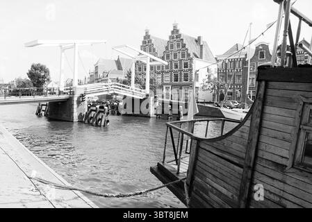Verschiedene Atmosphäre Haarlem the Netherlands, Whizgle News, Dutch Desk, Niederlande, 1950 - 2000 am 23-08-1995. Dies sind die Elemente im Bild. Die Szene zeigt eine ruhige Lage am Wasser, die von einer Mischung aus historischer Architektur und einer ruhigen Atmosphäre geprägt ist. Auf der linken Seite ist ein verwittertes Holzboot vertäut, dessen dunkle Holztöne sich vom sanften Licht der Umgebung abheben. Die Krümmungen und rustikalen Details des Bootes verleihen dem Boot ein Gefühl von Charakter und Nostalgie. Im Hintergrund überspannt eine malerische Zugbrücke den Wasserweg, deren weiße Struktur stark gegen die gedämpfte Seite gerichtet ist Stockfoto