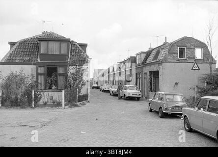 Häuser Amerika-buurt Hillegom Houses and Housing Construction, Whizgle News, Dutch Desk, Niederlande, 1950 - 2000 am 03-11-1977. Diese Themen sind in der Abbildung dargestellt. Die Szene zeigt eine ruhige Wohnstraße in einem älteren Viertel. Auf der linken Seite hebt sich ein zweistöckiges Haus mit einem schrägen, gekachelten Dach hervor, das sich durch eine Kombination aus dunklen und hellen Tönen auszeichnet. Die Fenster haben einfache Rahmen, eines mit einer kleinen, überhängenden Markise. Das Haus ist umgeben von grünen Blättern, die der Szene einen Hauch von Natur verleihen. Auf der anderen Straßenseite befindet sich eine Reihe einstöckiger Häuser, eac Stockfoto