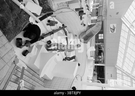 Arbeit an der Gran Dorado Folie, Whizgle News, Dutch Desk, Niederlande, 1950 - 2000 am 09-02-1992. Das Bild enthält diese Themen. Die Szene zeigt eine geschäftige Bau- oder Installationsstelle im Innenbereich, die durch einen großen, offenen Raum mit hellem, diffusem Tageslicht, das durch eine Glasdecke filtert, beleuchtet wird. Die Umgebung scheint ein modernes Gebäude mit klaren Linien und einer zeitgenössischen Ästhetik zu sein. Im Vordergrund sind zwei Personen aktiv an ihrer Arbeit beteiligt. Eine Person, die links positioniert ist, ist gehockt und konzentriert sich auf einen Behälter, während sie eine dunklere Flüssigkeit auf den aufträgt Stockfoto