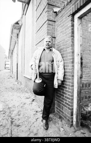 Herr Weijers Vorsitzender von Crescendo, Whizgle News, Dutch Desk, Niederlande, 1950 - 2000 am 27.03.1991. Das Bild enthält diese Themen. In einer düsteren urbanen Umgebung steht ein Mann vor einer Ziegelmauer, die verwitterten Charme ausstrahlt. Die Ziegelsteine in verschiedenen Rot- und Brauntönen bilden eine strukturierte Kulisse, die durch abblätternde Farbe und Alterserscheinungen akzentuiert wird. Der Boden ist mit losem Boden bedeckt, was auf eine kürzliche Baumaßnahme oder Renovierung hindeutet. Der Mann, der Selbstvertrauen ausstrahlt, hält eine Trompete sanft in der rechten Hand, während er seinen linken Fuß an die Wand legt. Er trägt eine helle Jacke, die einen Kontrast bildet Stockfoto
