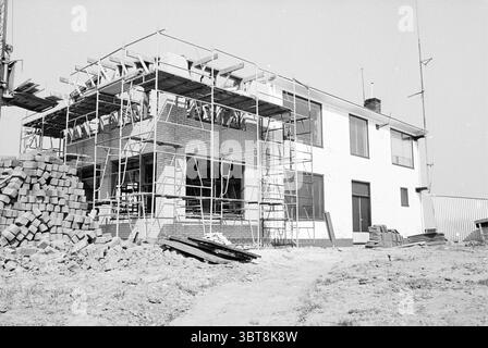 Renovierung des Vletterlieden Büros in IJmuiden Renovierungen IJmuiden Niederlande, Whizgle News, Dutch Desk, Niederlande, 1950 - 2000 am 15-04-1980. Dies sind die Elemente im Bild. Die Szene zeigt ein teilweise gebautes Gebäude, das sich durch ein schlichtes Äußere in den Farben weiß und Grau auszeichnet. Gerüste umrahmen die Struktur, was auf laufende Arbeiten hinweist. Das Gerüst besteht aus einem stark metallischen Grau, das Holzbalken und -Dielen trägt, die willkürlich über die oberen Ebenen angeordnet sind. Im Erdgeschoss sind lose Ziegelsteine sichtbar, die in organisierter Weise gestapelt sind, was darauf hindeutet Stockfoto
