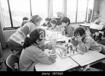Schulschach Wettbewerb Bavoschool Chess, Whizgle News, Dutch Desk, Niederlande, 1950 - 2000 am 13-01-1988. Diese Themen sind in der Abbildung dargestellt. Die Szene fängt ein hektisches Klassenzimmer mit Aktivität und konzentrierter Konzentration ein. Natürliches Licht strömt durch große Fenster und wirft sanfte Schatten über den Raum und schafft eine warme, einladende Atmosphäre. Im Vordergrund versammelt sich eine Gruppe von Kindern um mehrere Schachbretter, die auf ihren Tischen liegen. Die Schachfiguren, die sich zwischen dunklen und hellen Farben abwechseln, sind sorgfältig angeordnet, einige im Spiel mit Spielfiguren, die für den strategischen Mann ausgelegt sind Stockfoto