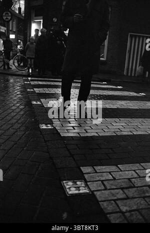 Beleuchtetes Zebra überquert Beverwijk Koningstraat Niederlande, Whizgle News, Dutch Desk, Niederlande, 1950 - 2000 am 16-11-1963. Diese Themen werden in der Abbildung angezeigt. Die Szene fängt eine geschäftige urbane Umgebung ein, die hauptsächlich durch künstliches Licht beleuchtet wird. Im Vordergrund steht ein Paar dunkler Schuhe am Rande eines Fußgängerübergangs, die die Höhe der Fußgängeransicht betonen. Der Pflastersteinpflaster reflektiert den Glanz des jüngsten Regens und schafft eine glänzende Oberfläche, die der Komposition Tiefe verleiht. Nebenbei dringt eine Gruppe von Menschen zusammen, die verschwommen, aber sichtbar sind, während sie darauf warten, die zu überqueren Stockfoto