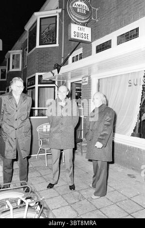 Gründer des Segelverbandes H.Z.V. Zeilen, Whizgle News, Dutch Desk, Niederlande, 1950 - 2000 am 09-12-1976. Dies sind die Elemente im Bild. In einer nächtlichen urbanen Umgebung stehen drei Männer vor einem Café mit großen Fenstern und geben einen Einblick in die gemütliche Inneneinrichtung. Das Gebäude verfügt über eine Ziegelfassade mit großen Glasscheiben, die das sanfte Leuchten der Lichter in der Nähe reflektieren. Oberhalb des Eingangs steht ein kreisförmiges Schild mit dem Namen einer Biermarke, während ein weiteres Schild den Namen des Cafés in einem gut beleuchteten Bereich anzeigt. Die drei Figuren tragen formelle Oberbekleidung: Zwei tragen ein langes, dunkles Over Stockfoto