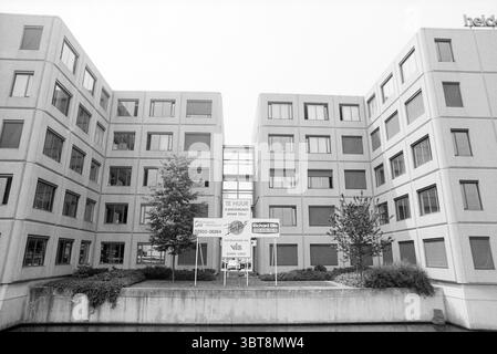 Büropark Beukenhorst Hoofddorp van Heuven Goedhartlaan Niederlande, Whizgle News, Dutch Desk, Niederlande, 1950 - 2000 am 24-05-1992. Dies sind die Themen im Bild. Die Szene zeigt eine moderne architektonische Umgebung, die durch eine Reihe rechteckiger, mehrstöckiger Gebäude gekennzeichnet ist, die um einen zentralen Innenhof angeordnet sind. Die Strukturen bestehen hauptsächlich aus einem gedämpften grauen Material, das eine zeitgenössische Ästhetik unterstreicht. Die Fassade weist ein sich wiederholendes Muster von Fenstern auf, die ordentlich eingerahmt sind und ein Gefühl der Gleichmäßigkeit widerspiegeln. Im Vordergrund ein paar kleine Bäume Stockfoto