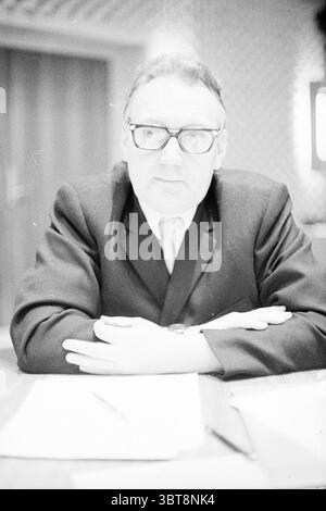 Beverwijk council leitet den Beverwijk gemeinderat, Whizgle News, Dutch Desk, Niederlande, 1950 - 2000 am 27-01-1972. Dies sind die Themen im Bild. Die Szene zeigt einen Mann, der an einem Tisch sitzt und ein Gefühl von Formalität und Autorität ausstrahlt. Er trägt einen dunklen Anzug, der klassisch und gut geschnitten wirkt, ergänzt durch eine helle Krawatte. Seine Arme sind auf dem Tisch vor ihm gefaltet, was auf eine nachdenkliche oder kontemplative Haltung hindeutet. Seine große, runde Brille unterstreicht sein ernstes Verhalten und umrahmt seinen fokussierten Ausdruck, der sowohl Vertrauen als auch Prüfung vermittelt. Der Hintergrund reve Stockfoto
