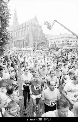 TROS laufen Start- und Ziellauf Haarlem Grote Markt the Netherlands, Whizgle News, Dutch Desk, Niederlande, 1950 - 2000 am 22. 10. 1988. Dies sind die Elemente im Bild. Die Szene strotzt vor Energie, ein lebhaftes Outdoor-Event, bei dem zahlreiche Teilnehmer auf den Straßen rasen. Eine große Menge von Läufern wird mitten im Schritt gefangen, viele tragen verschiedene sportliche Outfits und zeigen eine Mischung aus Farben, darunter helle Rot-, Blau- und Weißtöne, die von der monochromatischen Atmosphäre der Gesamtszene kontrastiert werden. Die Läufer zeigen eine Reihe von Ausdrücken, von Entschlossenheit bis hin zu Kameradschaft. In Stockfoto