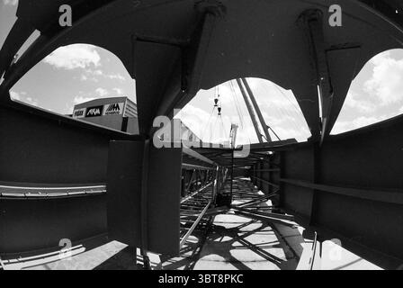 Crane at Figee, Whizgle News, Dutch Desk, Niederlande, 1950 - 2000 am 22-05-1991. Die Abbildung zeigt diese Themen. Die Szene präsentiert eine industrielle Umgebung, die durch Stahlträger in geometrischem Rahmen gekennzeichnet ist. Die Perspektive ist niedrig und abgewinkelt und erfasst das komplizierte Gitter von Metallarbeiten, die sich in Richtung eines zentralen Brennpunkts konvergieren. Die in einem gedämpften Grauton lackierten Balken weisen einen metallischen Glanz auf, der das Umgebungslicht reflektiert und ihr robustes Erscheinungsbild unterstreicht. Oben offenbart sich der Himmel durch die Lücken in der Struktur und zeigt ein helles azurblaues, von Grippe durchzogenes Licht Stockfoto