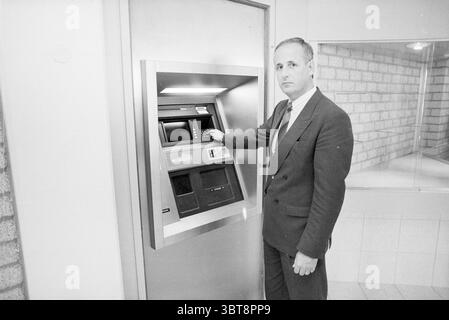 Porträt des Mannes verwendet Geldautomaten, Whizgle News, Dutch Desk, Niederlande, 1950 - 2000 am 06-1989. Diese Themen werden in der Abbildung angezeigt. In einer monochromen Umgebung steht ein Mann selbstbewusst in der Nähe eines Geldautomaten (ATM) aus Metall. Er trägt einen maßgeschneiderten dunklen Anzug, der ihm ein formelles Aussehen verleiht. Der Anzug steht im Kontrast zu den helleren Farben der Umgebung, die sich durch beige Ziegelwände und einen glatten, weißen Boden auszeichnet. Der Geldautomat ist elegant und modern, mit einer glänzenden silbernen Oberfläche, die das Licht im Raum reflektiert. Der Mann wird leicht zur Seite gedreht, in die Bedienung der Maschine eingegriffen, mit seinem R Stockfoto