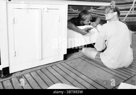 Segeltour mit dem Schoner Te Vega Scheveningen, Whizgle News, Dutch Desk, Niederlande, 1950 - 2000 am 15-08-1990. Dies sind die Themen im Bild. In dieser Szene sind zwei Personen in einem Moment auf einem Boot oder Schiff beschäftigt. Die Einrichtung verfügt über eine Holzoberfläche mit unterschiedlichen, gleichmäßig angeordneten Dielen, die den Boden bilden und zu einer nautischen Atmosphäre beitragen. Auf der linken Seite ist ein Schrank oder Fach teilweise geöffnet, was auf einen Bereich hindeutet, der für die Lagerung oder Ausrüstung ausgelegt ist. Eine Person hockt sich, blickt in den offenen Raum unter der Theke, ihre Hände greifen ein Gerät und konzentrieren sich intensiv Stockfoto