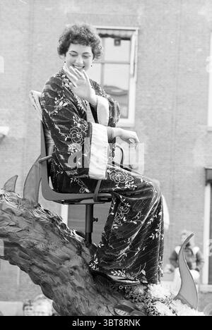 Tulip Queen auf KLM Parade Float Flower Parade Blumenausstellung, Whizgle News, Dutch Desk, Niederlande, 1950 - 2000 am 22-04-1988. Das Bild enthält diese Themen. In einer lebendigen Outdoor-Szene sitzt eine Person auf einem dekorativen Schwimmer und strahlt Freude und Selbstvertrauen aus. Die Person trägt ein elegantes Gewand mit komplizierten Blumenmustern, die tiefe Schwarztöne und leuchtende Farbtupfer in den Designs zeigen und ein harmonisches und lebendiges Erscheinungsbild schaffen. Ihr Haar ist in weichen Locken gestylt, was zu einer unbeschwerten Atmosphäre beiträgt. Um den Wagen herum ist eine Menge Zuschauer Stockfoto
