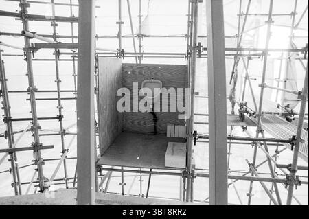 Verschiedene Baustellen Schiphol Schiphol, Whizgle News, Dutch Desk, Niederlande, 1950 - 2000 am 12-09-1989. Das Bild enthält diese Themen. Die Szene präsentiert eine Bauumgebung, die durch eine komplexe Anordnung von Gerüsten gekennzeichnet ist, wodurch eine gitterartige Struktur entsteht, die den Raum ausfüllt. Das Gerüst ist metallisch und verfügt über eine Kombination aus vertikalen und horizontalen Strahlen, von denen einige aufgrund des verfügbaren Lichts komplizierte Schatten erzeugen können. Im Zentrum der Komposition befindet sich ein kleines, rudimentäres Gehäuse aus Sperrholz, das einem funktionalen Zweck dient. Darin Stockfoto