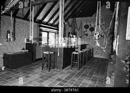 Holiday Farm Bakkum Farms, Whizgle News, Dutch Desk, Niederlande, 1950 - 2000 am 16-09-1975. Das Bild enthält diese Themen. Die Szene zeigt eine geräumige Küche mit rustikalem Charme. Die Wände sind mit freiliegenden roten Ziegeln verziert, was ein warmes und einladendes Ambiente schafft. Eine große zentrale Insel dominiert den Raum, aus dunklem Holz gefertigt und mit einer glatten, polierten Oberfläche, die einen schönen Kontrast zur Wärme der Ziegelsteine bildet. Rund um die Insel laden eine Reihe stabiler Holzhocker mit schlichtem Design zu zwanglosen Sitzgelegenheiten ein, die die einladende Atmosphäre der Küche noch verstärken. Natürliche Li Stockfoto
